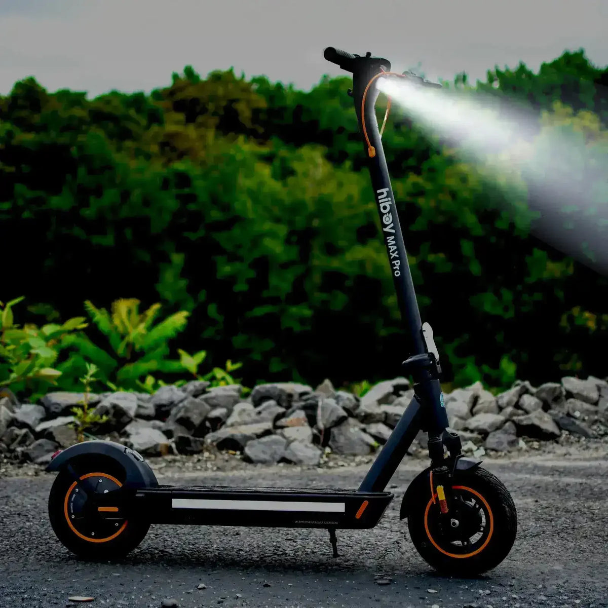 Electric scooter with headlight on, parked outdoors near rocks and greenery