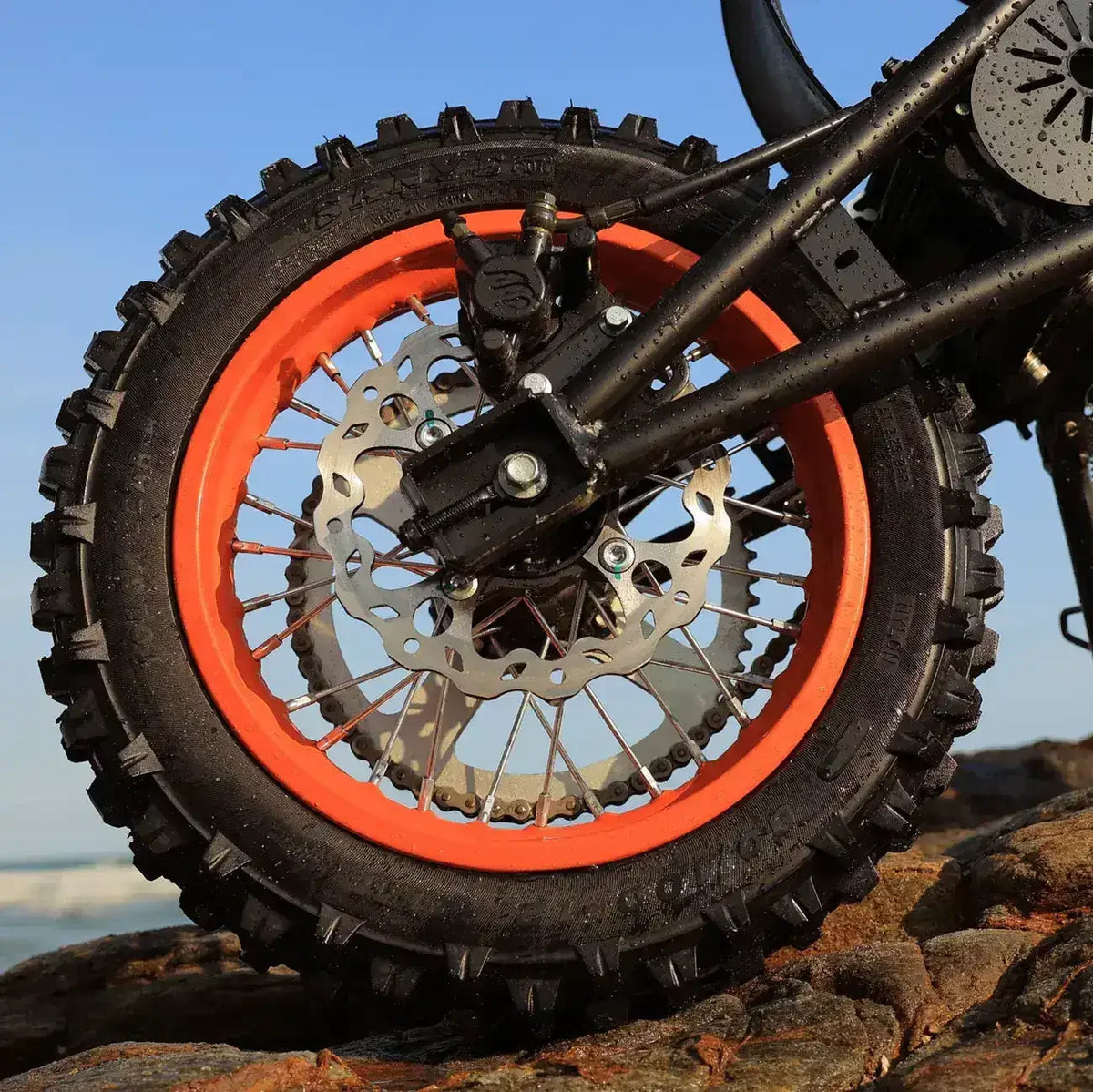 Close-up of a dirt bike rear wheel with orange rim and disc brake on rocky terrain.