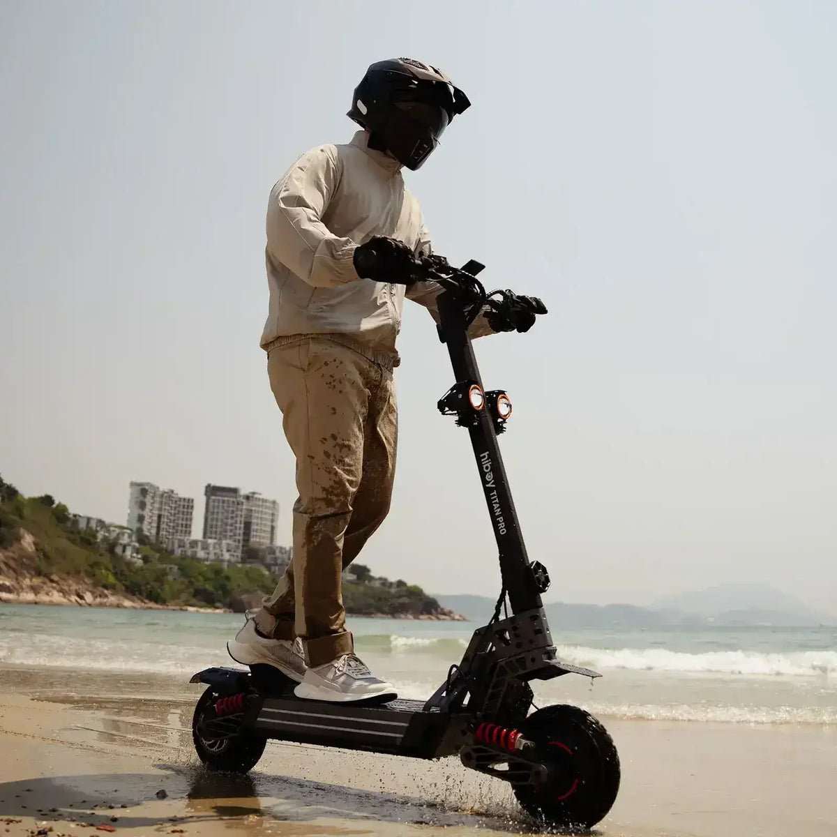 Person riding electric scooter on sandy beach near water, wearing helmet and gloves