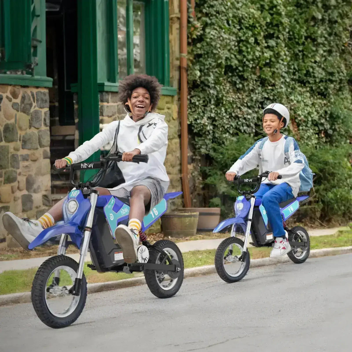 Two kids riding blue electric dirt bikes outdoors on a residential street, smiling and having fun.