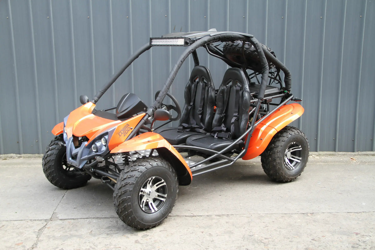 Orange two-seater go-kart with roll cage, off-road tires, and front headlights parked outside.
