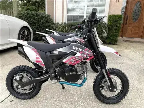 Two black and white kid dirt bikes with pink graphics parked on a driveway, knobby tires visible.
