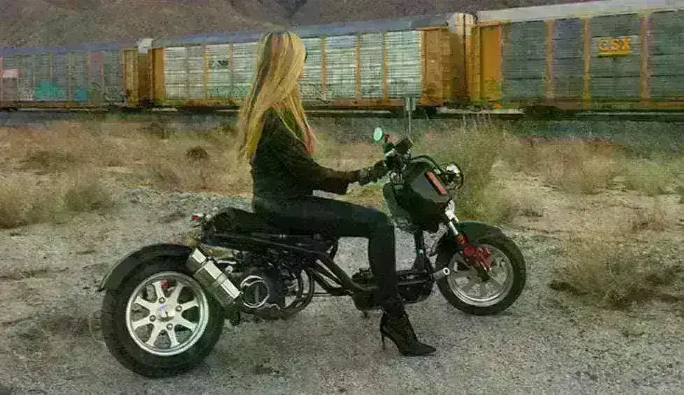Woman sitting on a black 50cc Maddog scooter outdoors near a train, desert landscape