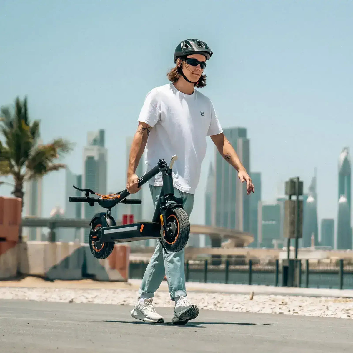 Man with helmet carrying foldable electric scooter outdoors, cityscape in background