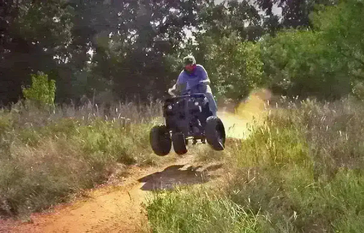 Helmeted rider jumping a dark Rhino utility ATV on a dirt trail between tall grass and trees