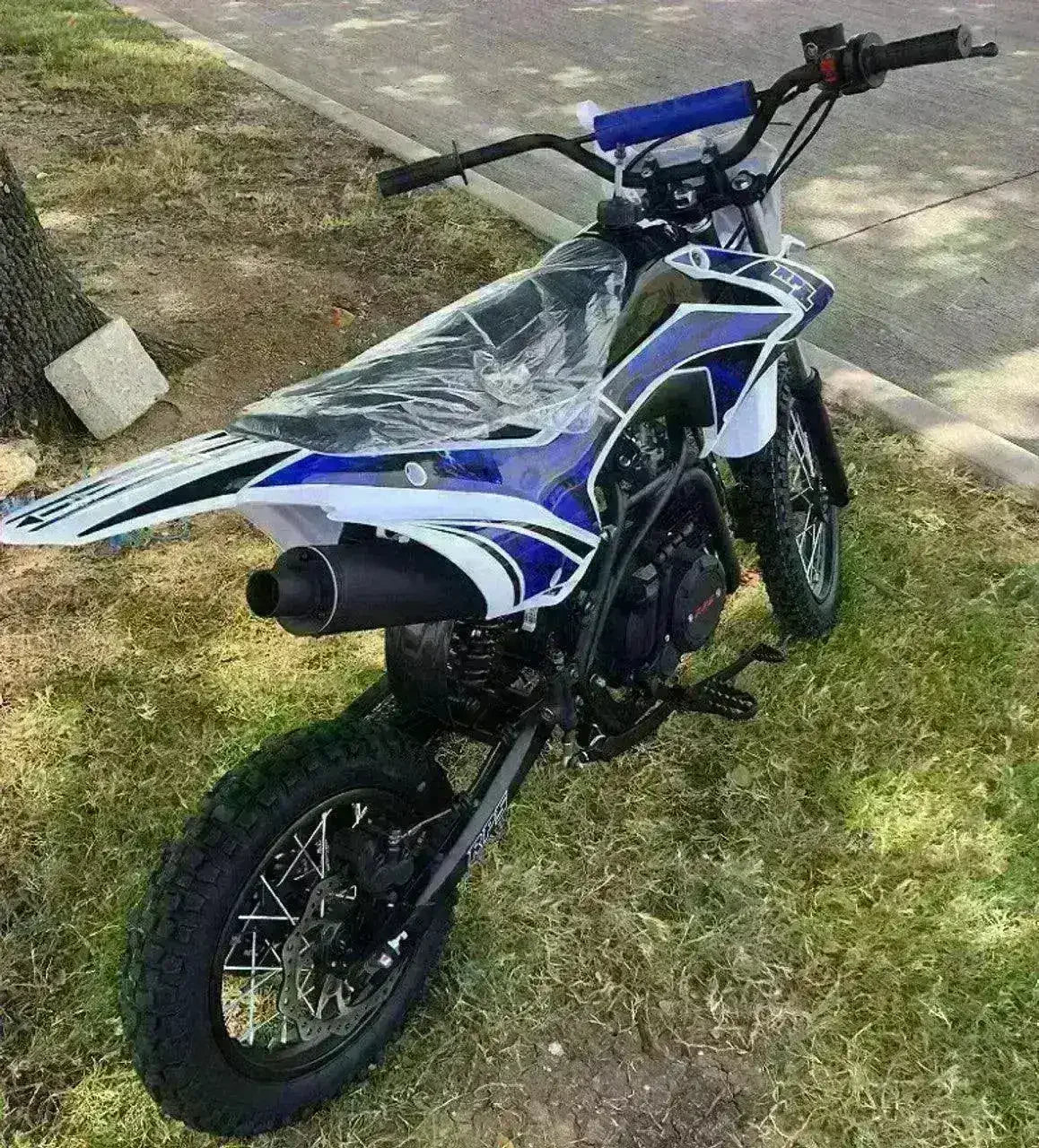 Blue and white dirt bike with off-road tires parked on grass near a sidewalk
