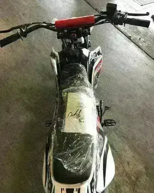 Top view of a youth dirt bike with black seat and red handlebars, on a concrete floor.
