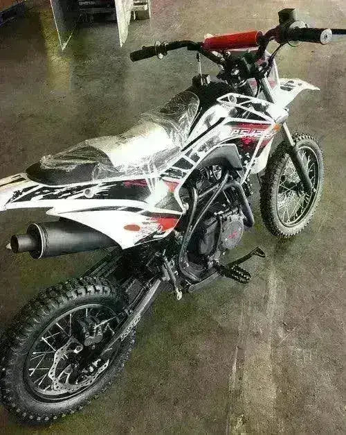 Rear three-quarter view of a white RPS 125S dirt bike showing exhaust, rear disc brake and plastic-covered seat indoors