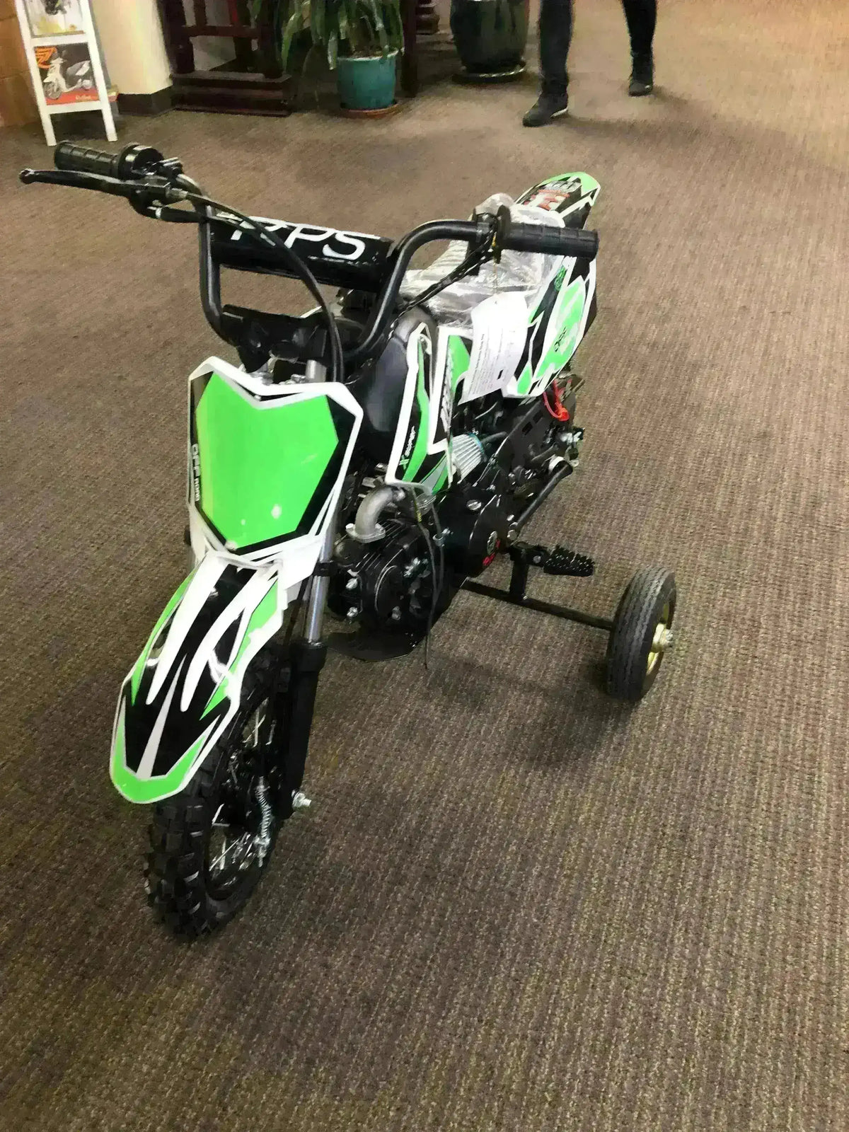 Green and white kids 70cc dirt bike with training wheels on indoor carpet