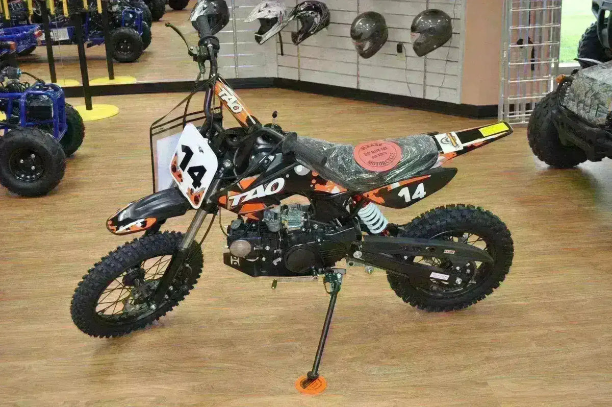 110cc youth dirt bike with black and orange graphics, displayed in showroom with helmets on wall