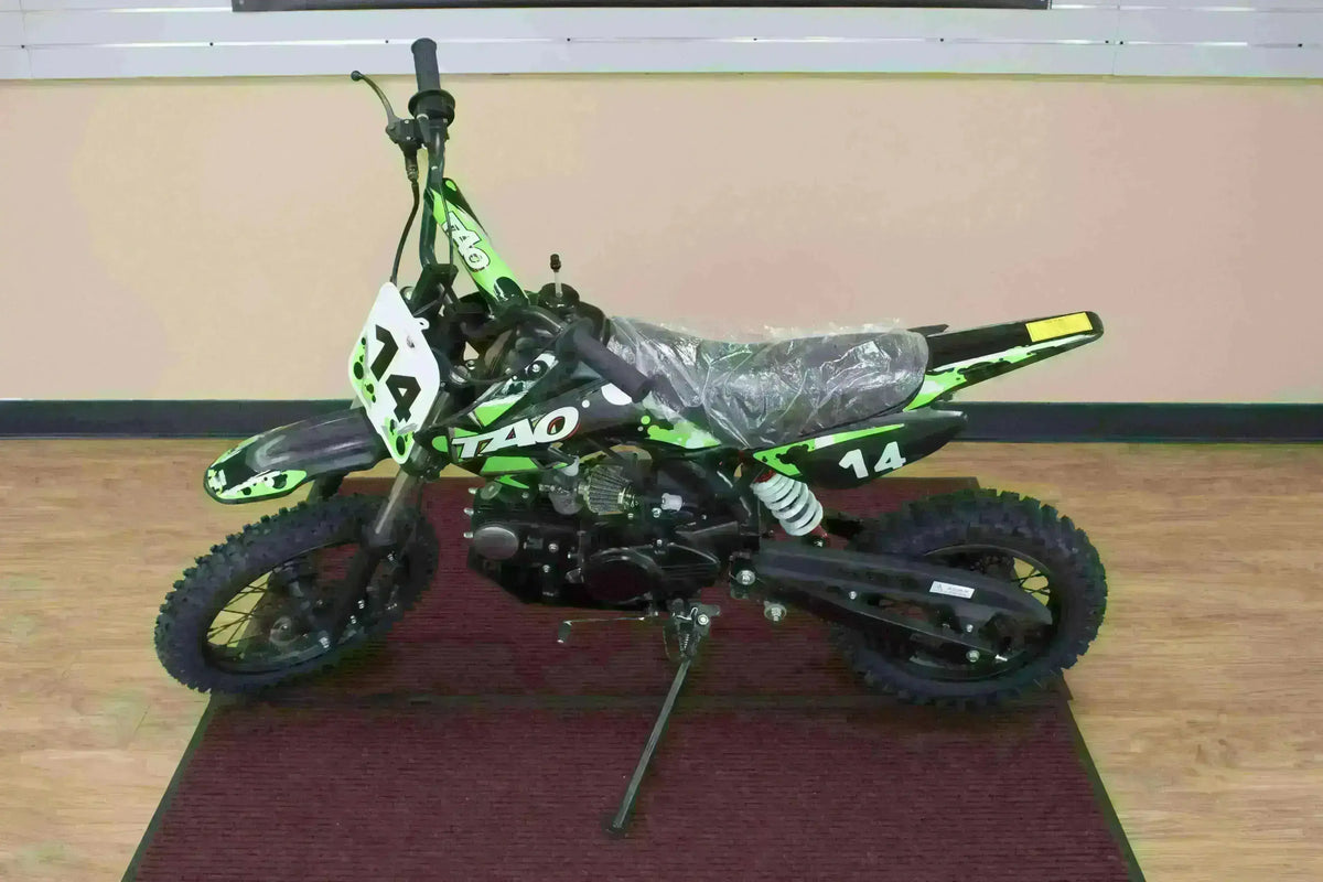 Green 110cc youth dirt bike with number 14, off-road tires, and side stand indoors