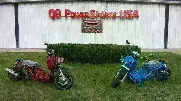 Red and blue 50cc scooters parked on grass in front of PowerSports storefront.