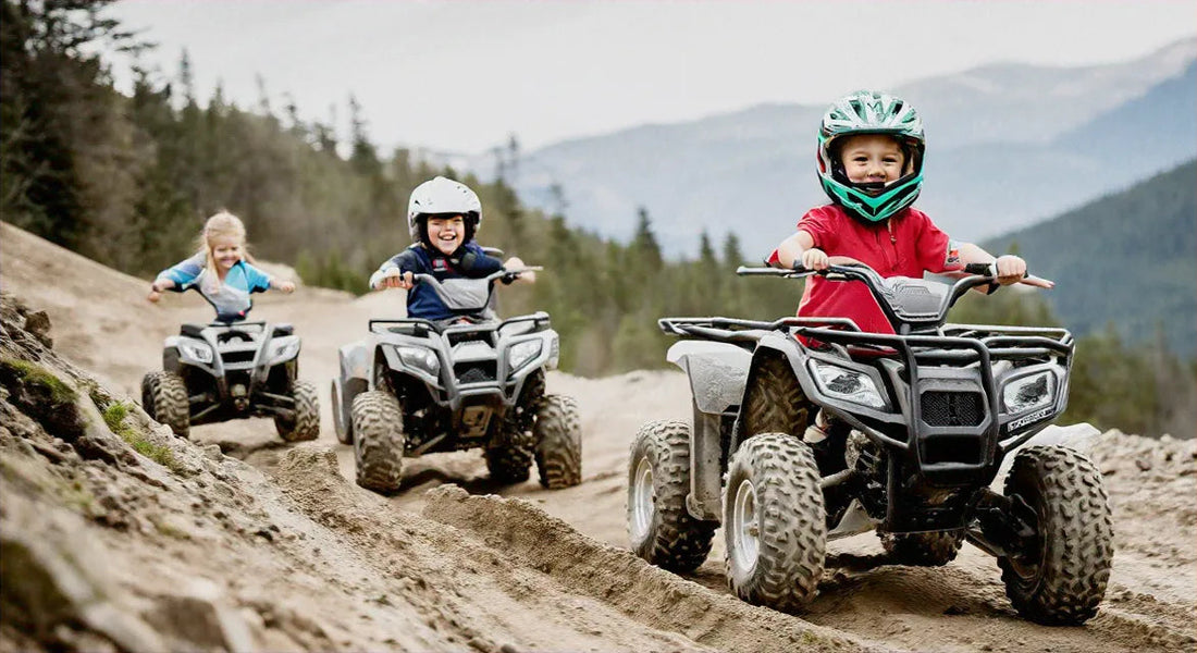 Kids riding youth ATVs on a dirt trail outdoors with forest and mountains in the background