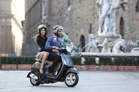 Two people riding a 50cc scooter in a city square, both wearing helmets.