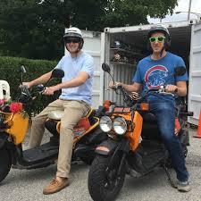 Two men wearing helmets riding orange 50cc scooters outdoors near a storage container
