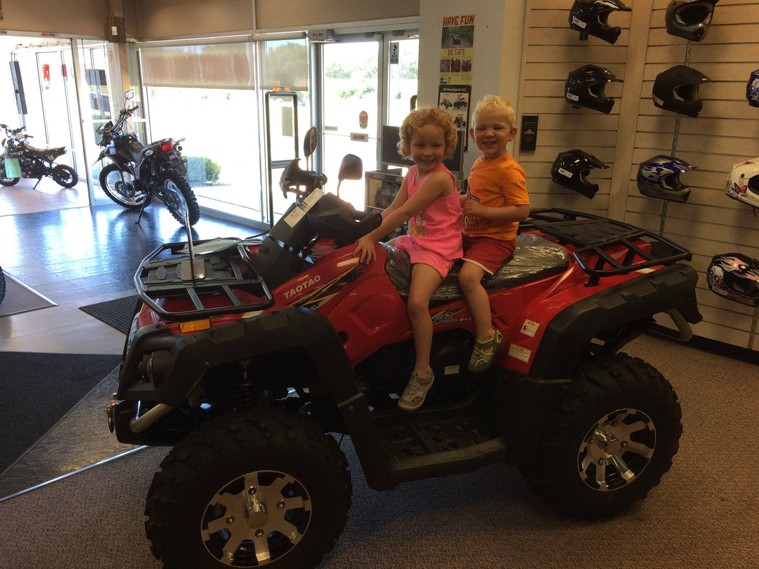 Two children sitting on a red ATV in a showroom with helmets and motorcycles on display.