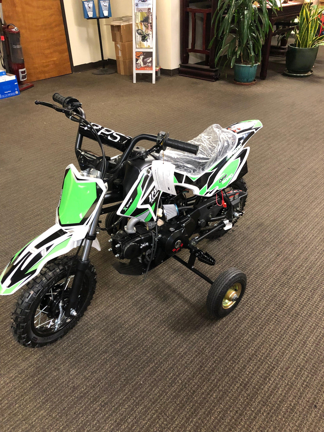 Green kids dirt bike with training wheels for beginners displayed indoors