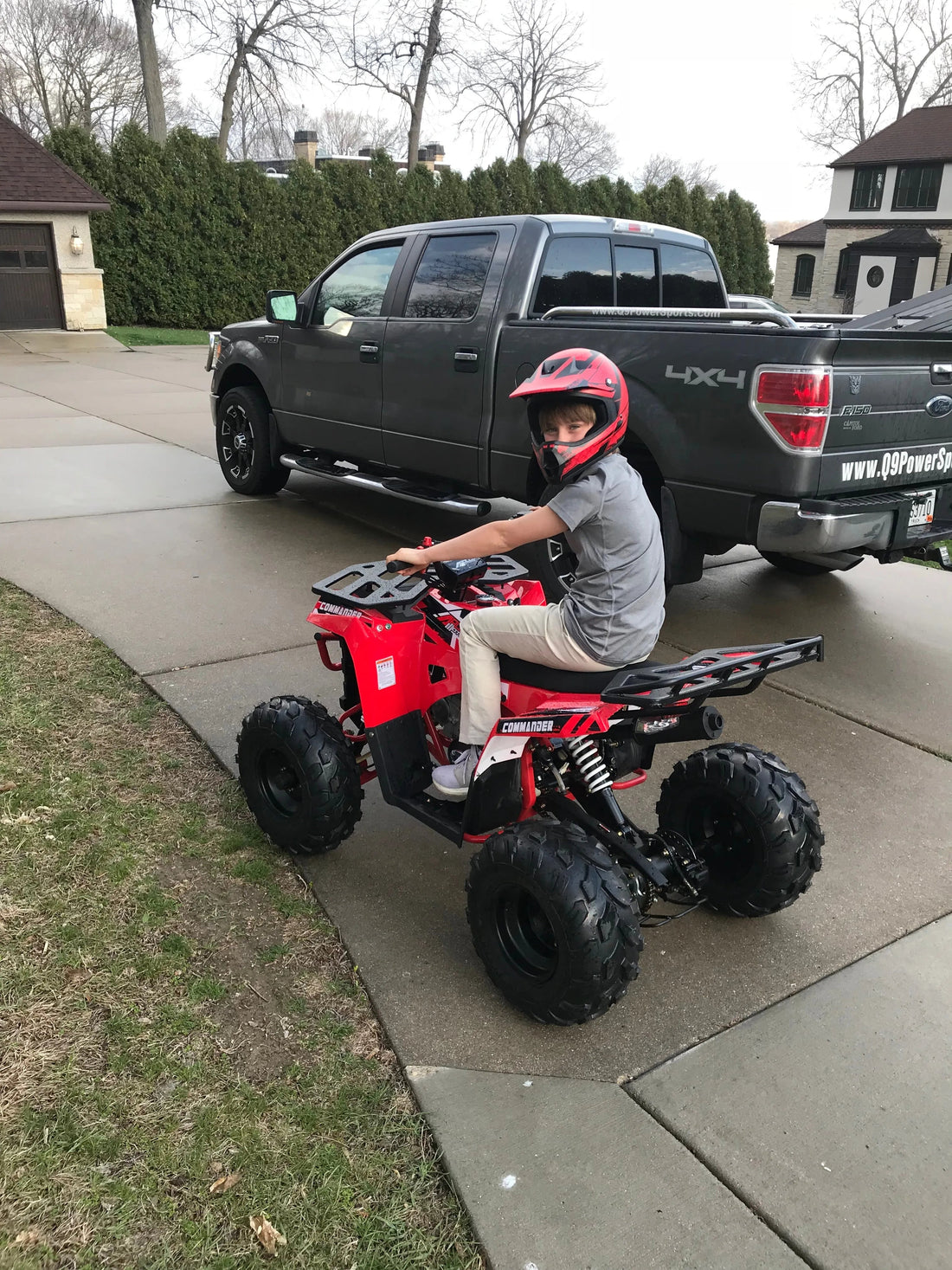 Child wearing red helmet riding a red youth ATV on driveway in front of black pickup truck and suburban homes