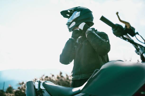 Rider in black gear adjusting helmet next to a dirt bike outdoors, clear sky in background