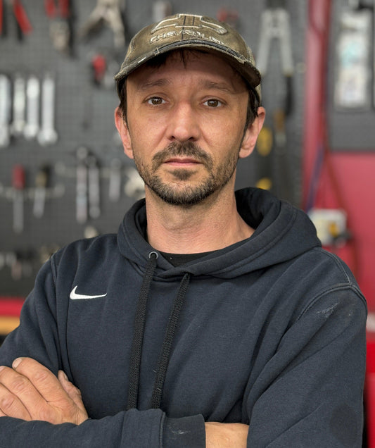 Mechanic in a workshop wearing a hoodie and cap, with tools hanging on the wall behind him.
