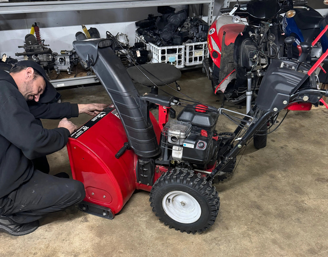 Mechanic repairing a red snow blower in a workshop with ATVs, dirt bikes, and scooters nearby.
