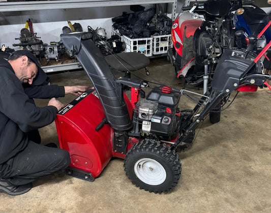 Mechanic repairing a red snow blower in a workshop with ATVs, dirt bikes, and scooters nearby.