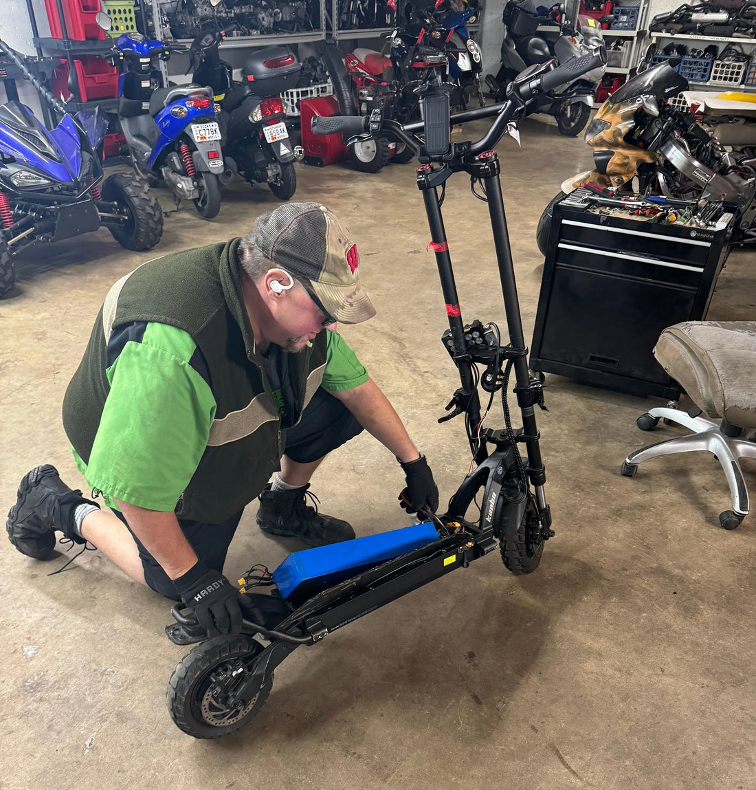 Technician repairing electric scooter battery in workshop with ATVs and scooters in background
