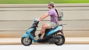Adult riding a blue scooter on a city street, wearing casual clothes and a backpack