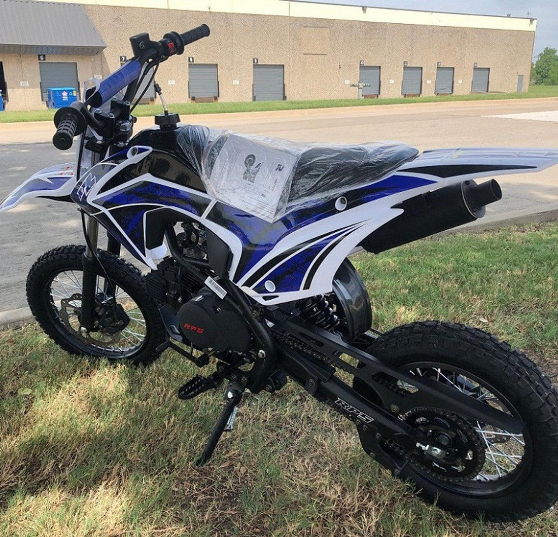 Blue and white youth dirt bike with plastic-covered seat parked on grass near industrial building