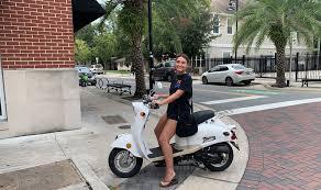 Young woman sitting on a white scooter parked on a city street near palm trees and buildings.