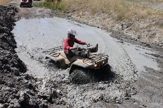 Rider wearing helmet and red shirt drives ATV through deep mud on outdoor off-road trail