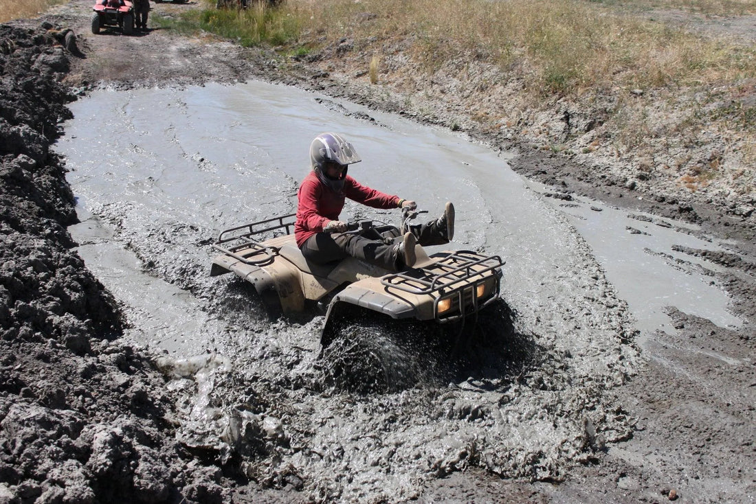 Person riding ATV through muddy trail with water splash, wearing helmet and red shirt