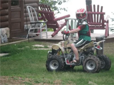 Child riding a small ATV on grass near a cabin, wearing a helmet and summer clothes.