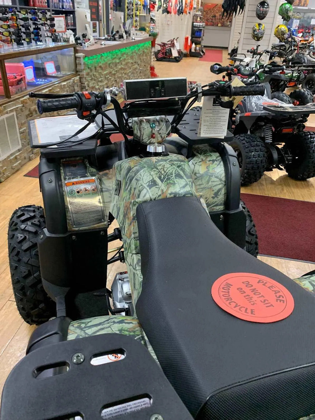 Camo ATV on showroom floor with a 'do not sit' sign, surrounded by helmets and other off-road vehicles.