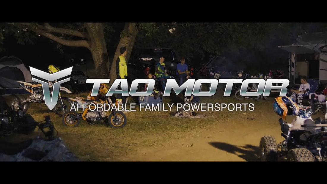 Group of people with dirt bikes, ATVs, and gear outdoors under trees at dusk
