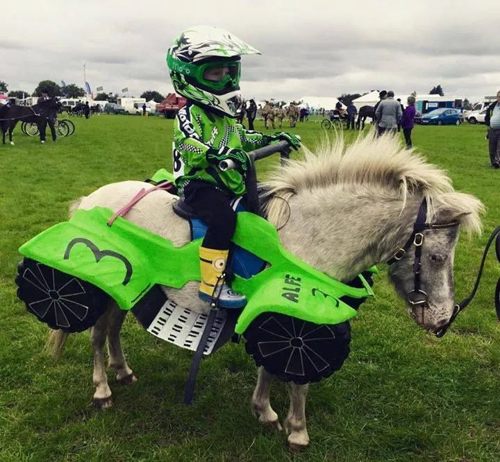Child in motocross gear riding a pony decorated to look like a green ATV at an outdoor event