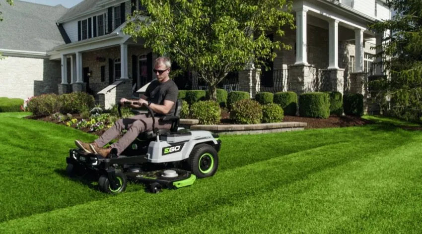 Man riding a zero turn lawn mower on a well-manicured lawn in front of a suburban home