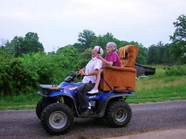 Two people riding a blue ATV with an armchair attached on a rural road, green trees in background.