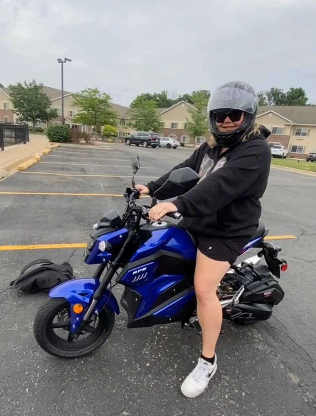 Person wearing helmet and sunglasses riding a blue mini motorcycle scooter in a parking lot