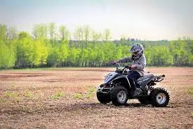 Person riding a black ATV on a dirt field with trees in the background