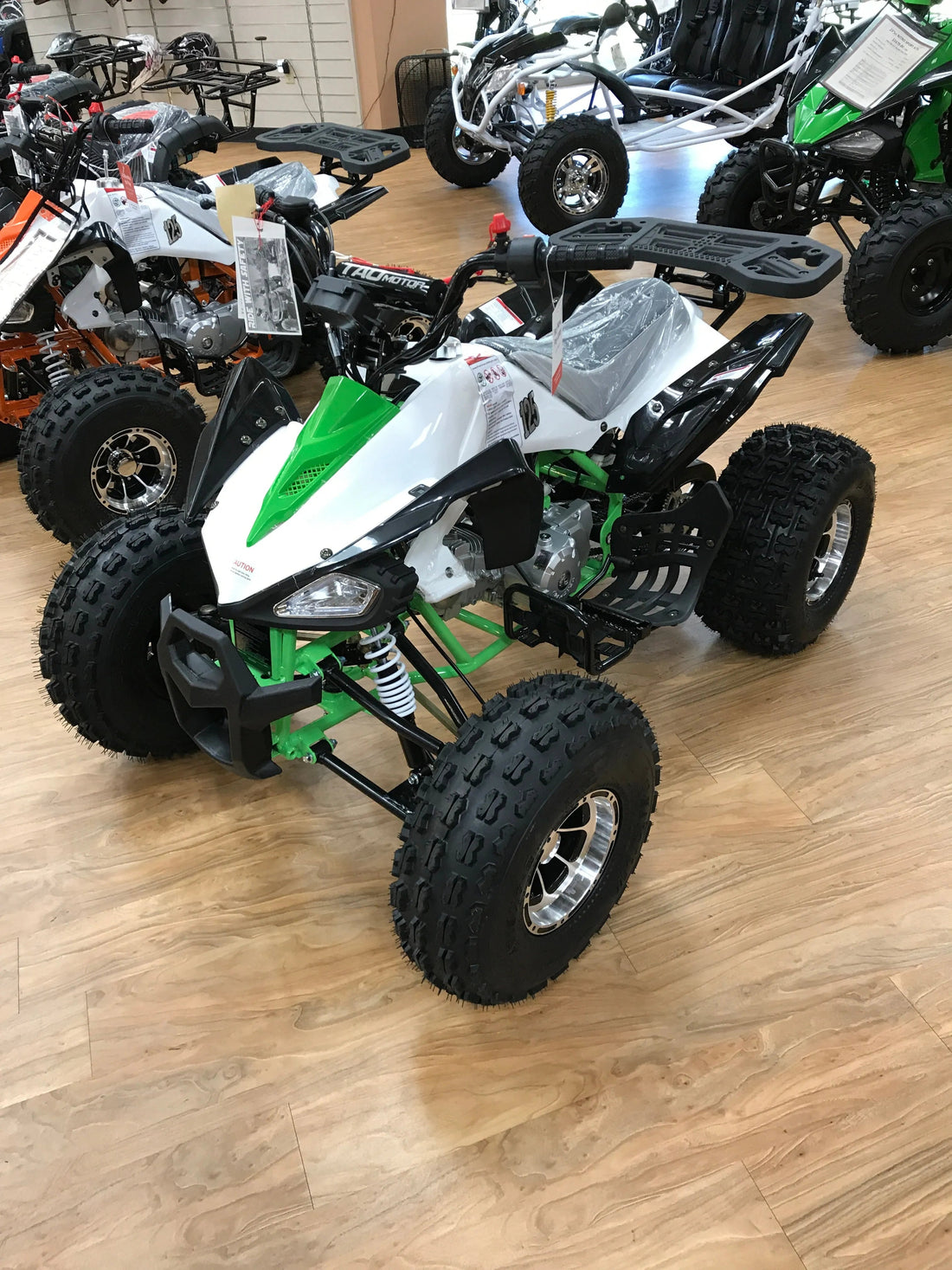Green and white ATV with off-road tires on display in a showroom with other powersports vehicles.
