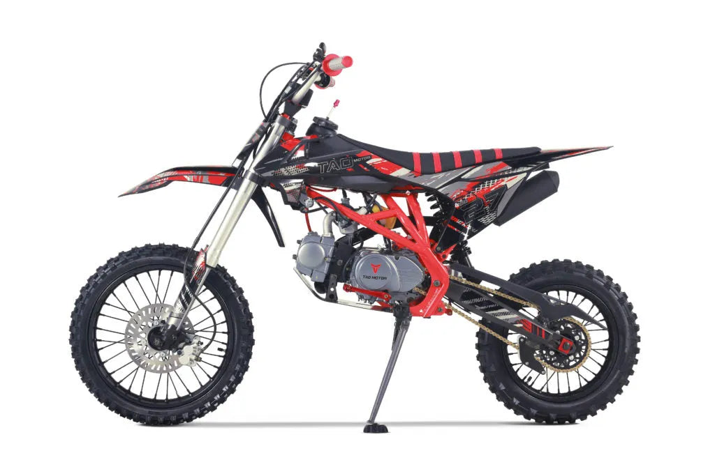 Side view of a red and black Taq dirt bike with knobby tires and a kickstand on a white background