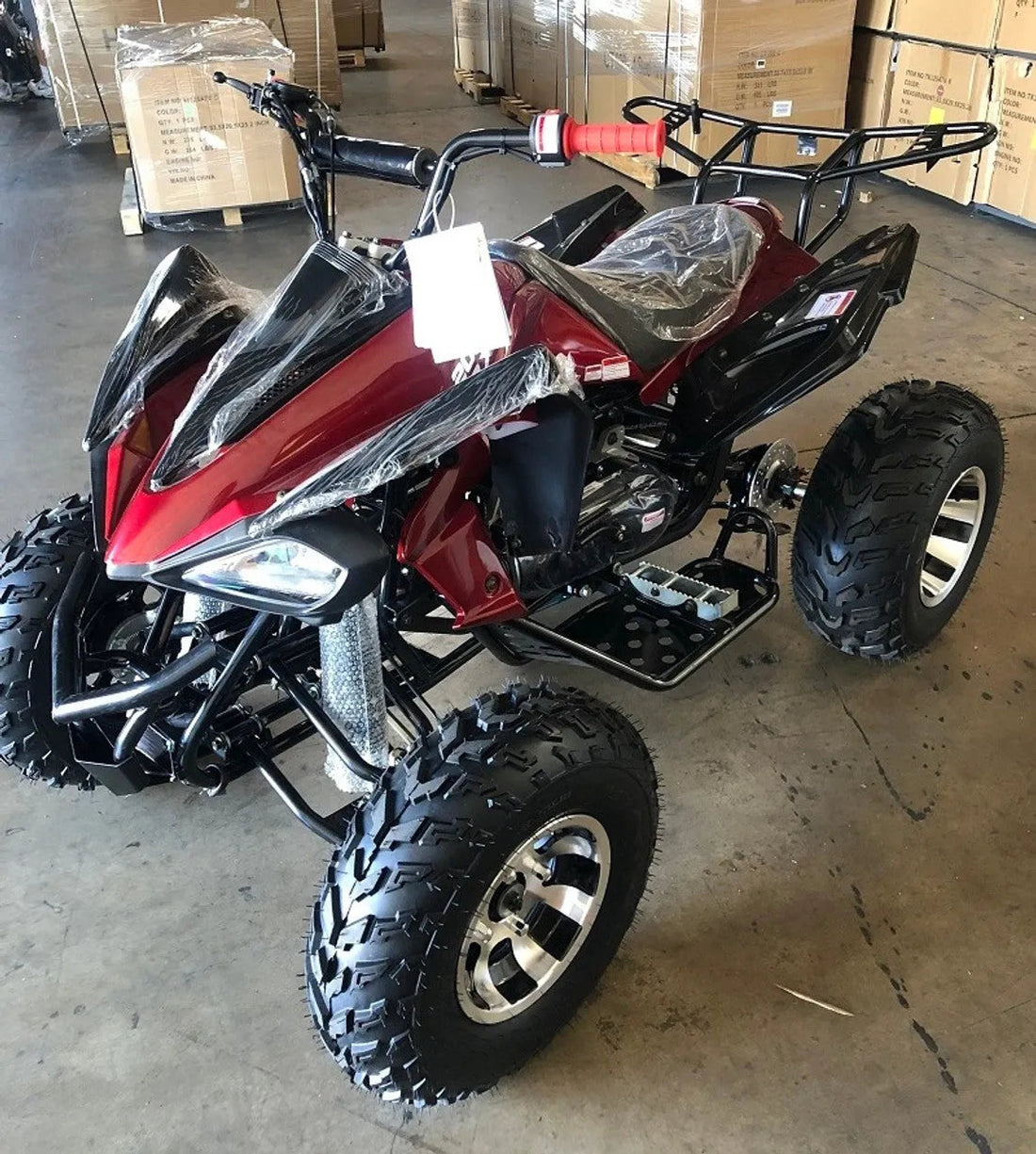 Red youth ATV with black frame and large knobby tires in warehouse with cardboard boxes