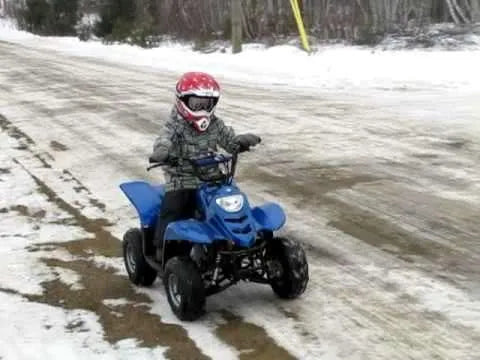 Child riding a blue ATV on a snowy, dirt road while wearing a red helmet and goggles