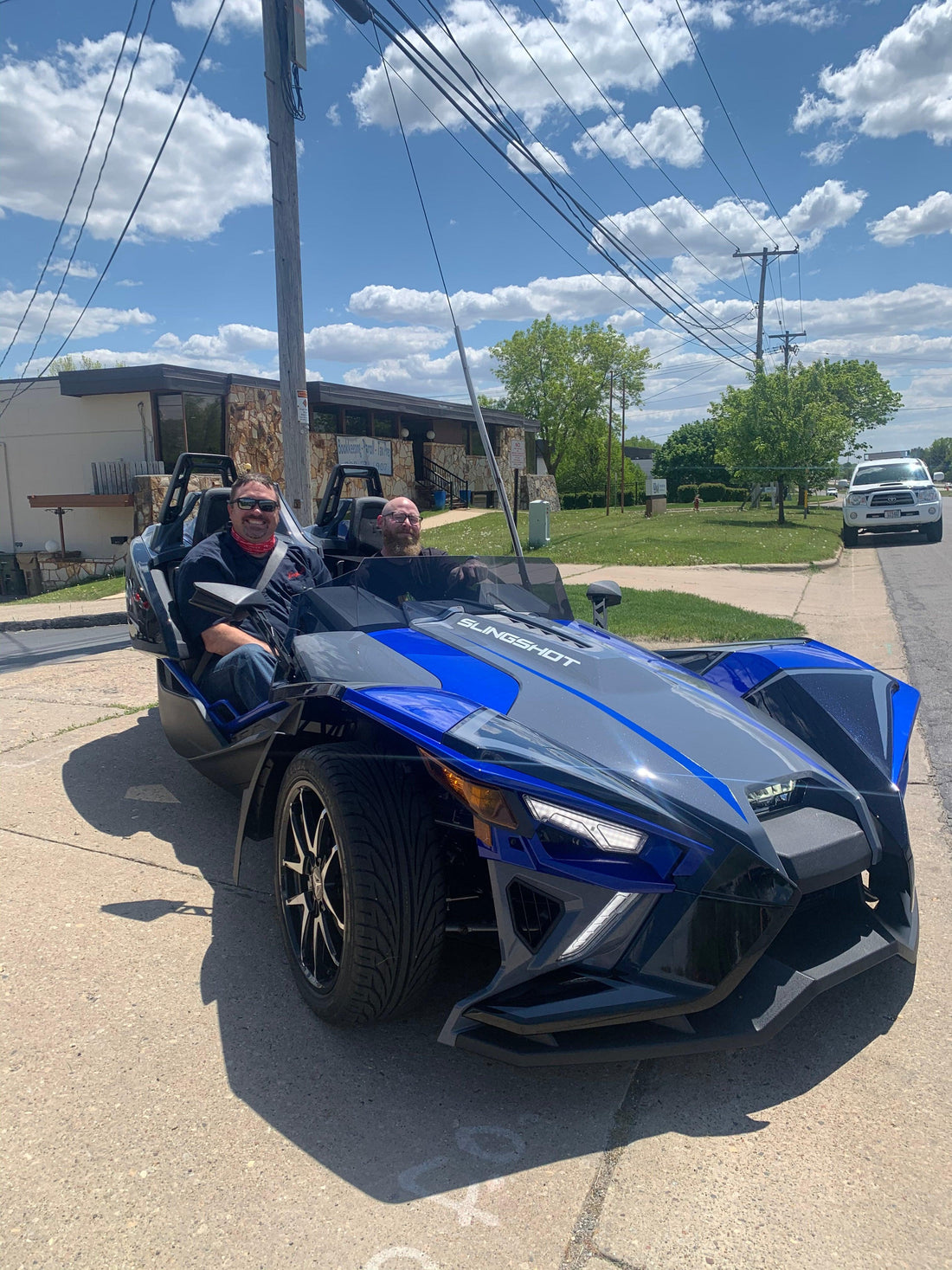 Two men in a blue Polaris Slingshot three-wheel vehicle parked on a sunny day in front of a building, perfect for spring powersports rides in southern Wisconsin.