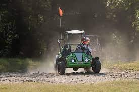Youth riding green gas-powered go-kart on dirt trail outdoors with safety flag.
