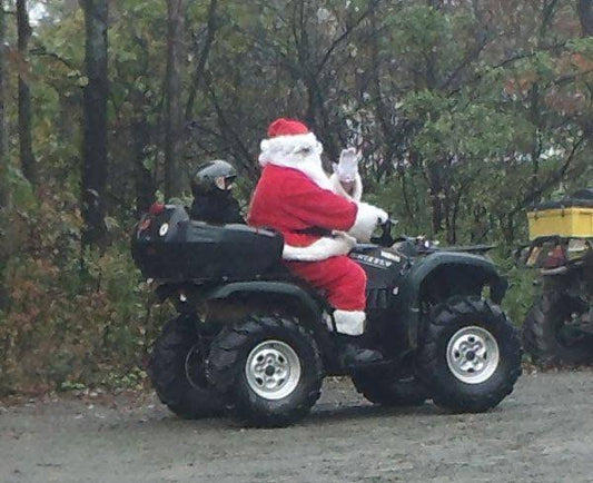 Santa Claus riding an ATV with a passenger in a forested outdoor setting