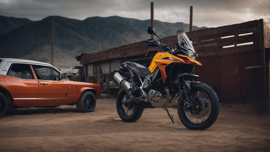 Orange adventure motorcycle and classic car parked outdoors on a dirt lot near mountains