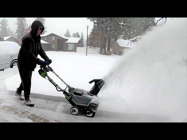 Person using a cordless snow blower to clear a snowy driveway in a residential neighborhood.