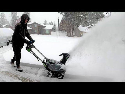 Person using a cordless snow blower to clear a snowy driveway in a residential neighborhood.